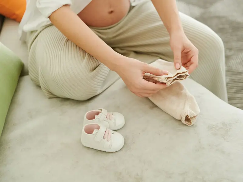 Une femme enceinte plie de tout petits vêtements à côté d'une paire de chaussons de bébé